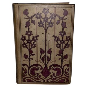May include: A vintage book with a tan cover and a burgundy floral design. The cover features a symmetrical pattern of stylized flowers and vines, with vertical lines. The book's spine is visible on the left side.