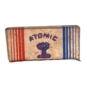 Op de afbeelding: Een rechthoekige doos met een bruine papieren wikkel. De wikkel heeft rode verticale strepen aan de linkerkant en blauwe verticale strepen aan de rechterkant. Het woord "ATOMIC" is blauw gedrukt, boven een blauwe en rode paddenstoelwolk-afbeelding.