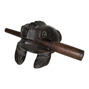Puede incluir: Un instrumento musical de madera de color marrón oscuro con forma de rana y un palo de madera. La rana tiene detalles tallados y un agujero para que pase el palo. El instrumento está diseñado para tocarse raspando el palo por la espalda de la rana.