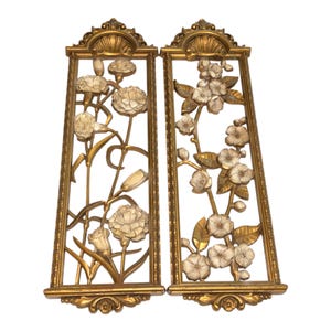 May include: A pair of rectangular wall plaques featuring gold-toned frames with ornate shell-like carvings at the top and bottom. The interior panels display raised, three-dimensional floral arrangements with white petals and gold stems and leaves.