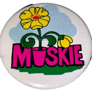 May include: A white badge featuring a yellow flower with a red centre and the word "MUSKIE" in pink. The design includes a green stem, a pink pot, and a blue and white cloud background.