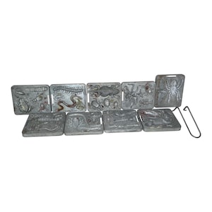 May include: A collection of nine rectangular metal casting molds featuring relief designs of various animals, insects, and sea creatures, including a frog, snake, spider, and lizard. A single metal handle tool is positioned to the right side.