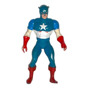 May include: A vintage action figure of a superhero in a blue and white costume with a star emblem. The figure has red gloves and boots. The head is blue with a mask and bear ears.