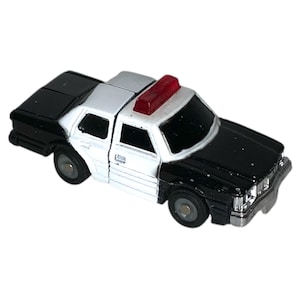 May include: A small police car toy. The toy car is black and white with a silver front bumper and a red light bar on the roof. The toy has four small wheels and is a classic toy car.