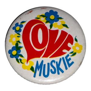 May include: A round, white button with a red heart-shaped design that says "LOVE" in white letters. Below the heart, the word "MUSKIE" is written in blue. The button is decorated with blue and yellow flowers.