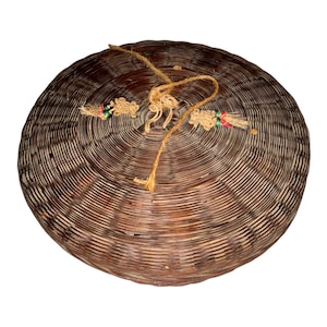 May include: A round, woven basket with a lid. The basket is made of brown and tan materials, with a spiral design. A tan string is attached to the lid, and there are decorative tassels with red, green, and white accents.