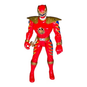 May include: A red action figure of a Power Ranger with gold and blue accents. The figure has a helmet, shoulder pads, and a belt. The figure is standing upright with its arms at its sides.