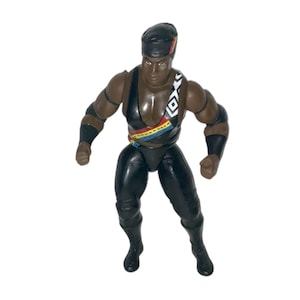 May include: A vintage action figure of a muscular man in a black outfit. The figure has a black cap, wristbands, and boots. The outfit features a colorful geometric design across the chest. The figure is posed with arms slightly outstretched.