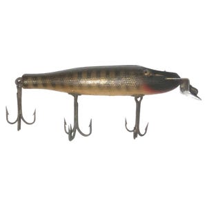 Puede incluir: Un señuelo de pesca vintage con un cuerpo dorado y negro, con tres anzuelos triples y una pequeña aleta metálica. El señuelo tiene un acento rojo cerca de la cabeza y está diseñado para imitar a un pez pequeño.