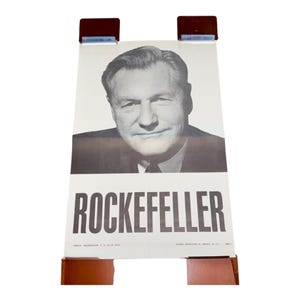May include: A black and white poster featuring a portrait of a man with the name "ROCKEFELLER" in bold letters. The poster is held by two brown wooden supports at the top and bottom.