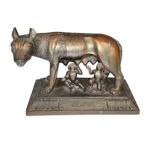 May include: Bronze sculpture of the Capitoline Wolf nursing Romulus and Remus. The sculpture is on a rectangular base with the word "ROMA" inscribed. The wolf and the twins are detailed with a bronze finish.