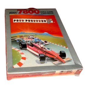 May include: Atari 7800 Pole Position II video game cartridge box. The rectangular gray box features a red and orange sunset background with a red race car driving on a track. The text reads Atari 7800 Video Game Cartridge and Pole Position II.