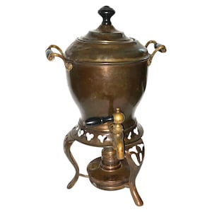 Puede incluir: Antigua cafetera de bronce con tapa y grifo. La cafetera tiene un asa negra en el grifo y la tapa, y dos asas a los lados. Se apoya sobre una base decorativa con tres patas.