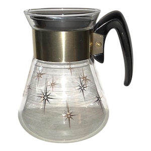 May include: Vintage glass pitcher with a black handle and gold-colored metal accents. The clear glass body features a pattern of starburst designs. The pitcher has a wide base and a narrow top, with a black lid.