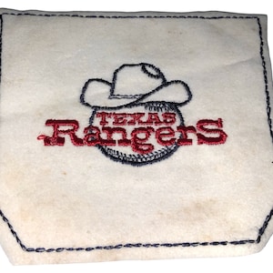 May include: Cream-colored square coaster with a stitched navy blue border. The center features a baseball with a cowboy hat above it and the words "TEXAS RANGERS" embroidered in red.