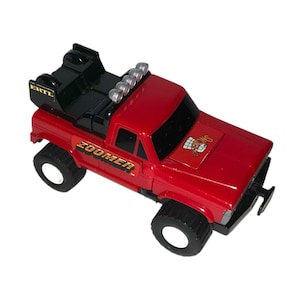 May include: A red toy truck with black tyres and a black tow hitch. The truck has the word "ZOOMER" printed on the side and a logo on the bonnet. The truck has a row of silver lights on the roof.