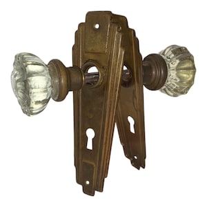 以下が含まれることがあります： 透明なガラス製のドアノブが2つ付いた、アンティークドアハードウェアセット。ノブは、鍵穴が付いた真鍮色の金属製バックプレートに取り付けられています。このハードウェアは、ヴィンテージのアールデコスタイルです。