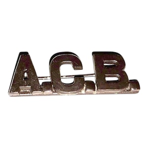 May include: A silver-toned metal pin featuring the letters "A.C.B." in a blocky font. The letters are connected and mounted on a horizontal bar with a pin clasp on the back. The pin is likely a vintage or custom accessory.