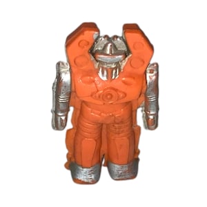 May include: A small, orange and silver robot toy. The toy has a humanoid shape with a detailed design, including a head, torso, arms, and legs. The robot's body is primarily orange, with silver accents on the head, arms, and feet. The toy is standing upright.