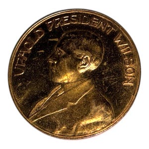 Op de afbeelding: Een goudkleurige munt met een profiel van een man in een pak. De rand van de munt heeft de woorden "UPHOLD PRESIDENT WILSON" in hoofdletters. De munt heeft een licht versleten uiterlijk.