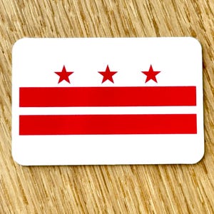 May include: A rectangular white sticker featuring the flag of Washington, D.C. The flag design includes three red stars above two horizontal red stripes.