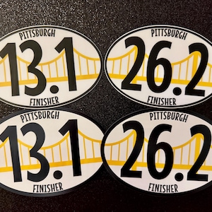 May include: Four oval-shaped stickers with a white background and black text. The stickers feature the text "Pittsburgh" and "Finisher" along with the numbers "13.1" and "26.2". A yellow bridge graphic is behind the numbers.