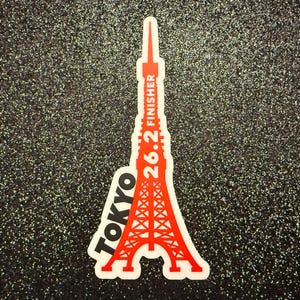 Puede incluir: Una pegatina blanca con un diseño de la Torre de Tokio roja. La torre está delineada en negro y tiene el texto "26.2 FINISHER" en el lateral. El texto "TOKYO" está escrito en diagonal en la parte inferior izquierda de la pegatina.