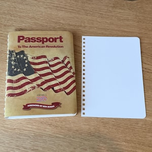 May include: A spiral-bound notebook and a small book with the title "Passport To The American Revolution". The book cover features a distressed American flag design in red, white, and blue. The notebook has blank white pages.