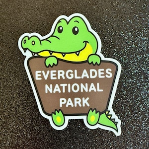 May include: A green cartoon alligator holding a brown sign that says "Everglades National Park".