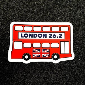 Może przedstawiać: Naklejka z czerwonego autobusu piętrowego z nadrukiem "LONDON 26.2" na boku i flagą Unii Jack na przodzie.