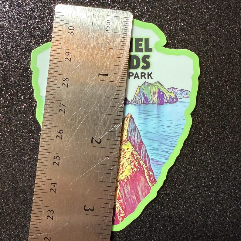 May include: A green and white sticker with a colorful illustration of a landscape. The sticker is shaped like a mountain range and has the text "Channel Islands National Park" on it. The sticker is approximately 3 inches tall.