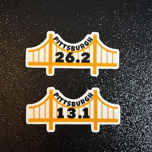 Puede incluir: Dos pegatinas blancas y amarillas con un diseño de puente. La pegatina superior dice "Pittsburgh 26.2" y la inferior "Pittsburgh 13.1". Sobre un fondo negro brillante.