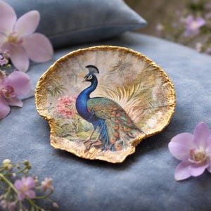 May include: Decorative, gold-edged trinket dish featuring a vibrant peacock illustration. The peacock is depicted with blue, green, and brown feathers, set against a backdrop of pink flowers and foliage. The dish has an irregular shape.