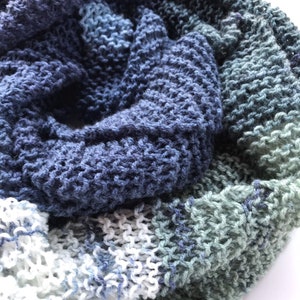 May include: A hand-knitted scarf in shades of blue, gray, and white. The scarf features a textured, open-knit design, with a gradient effect from dark blue to light gray and white. The soft, cozy scarf is perfect for cooler weather.