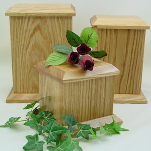 May include: Three wooden boxes with lids, made of light brown oak wood. The boxes are different sizes and are arranged in a staggered formation. The smallest box is in the front and the largest box is in the back. A sprig of green ivy and three red roses are in front of the boxes.
