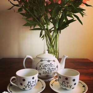 May include: A white ceramic teapot and two teacups with saucers. The teapot is decorated with the text "Married 10th June 2017" and floral accents. The teacups are labeled "Mrs. Viney" and have floral designs.