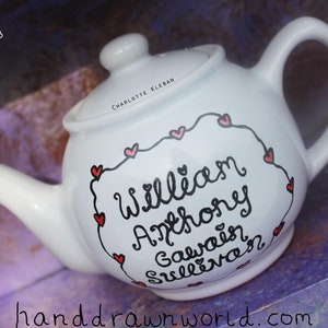 May include: White teapot with a black handwritten inscription that reads "William Anthony Galway Sullivan" surrounded by a black border with red hearts. The teapot is on a purple surface.