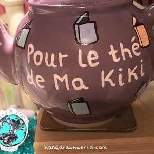May include: Purple teapot with white lettering that says "Pour le the de Ma Kiki". The teapot is decorated with hand-painted book illustrations.