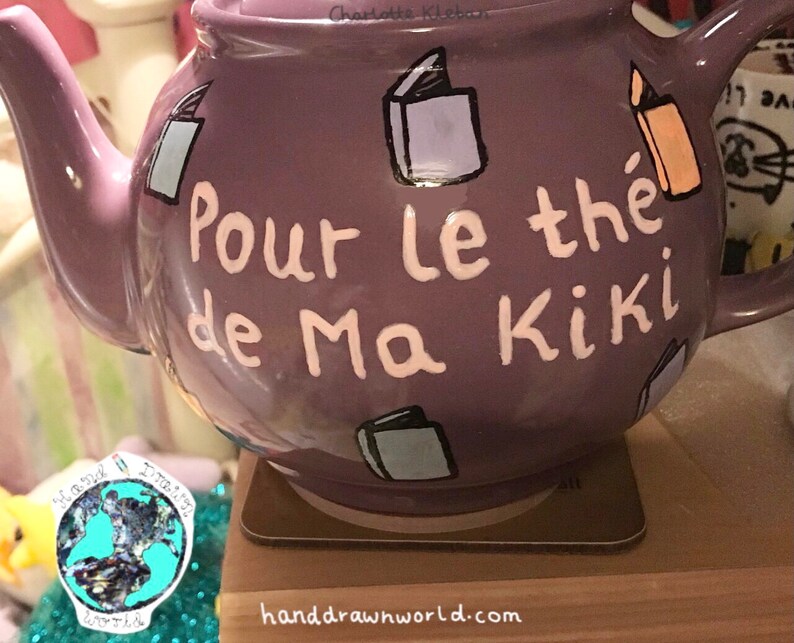 May include: Purple teapot with white lettering that says "Pour le the de Ma Kiki". The teapot is decorated with hand-painted book illustrations.