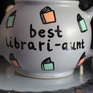 May include: A grey teapot with a black inscription that reads "best librari-aunt". The teapot is decorated with colourful book illustrations.