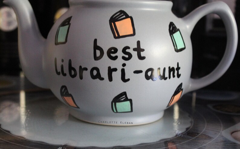 May include: A grey teapot with a black inscription that reads "best librari-aunt". The teapot is decorated with colourful book illustrations.