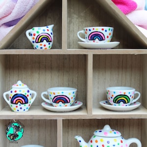 May include: A rainbow-themed tea set featuring a teapot, creamer, sugar bowl, and four teacups and saucers. The set is decorated with white polka dots and a rainbow with pink, orange, yellow, green, blue, and purple stripes. The teapot has the words "Felicity Rose" written on it.