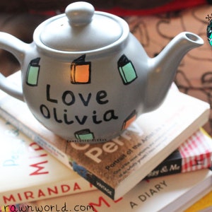 May include: A grey teapot with a white lid and black lettering that says "Love Olivia". The teapot is decorated with colourful book illustrations. The teapot is sitting on a stack of books.