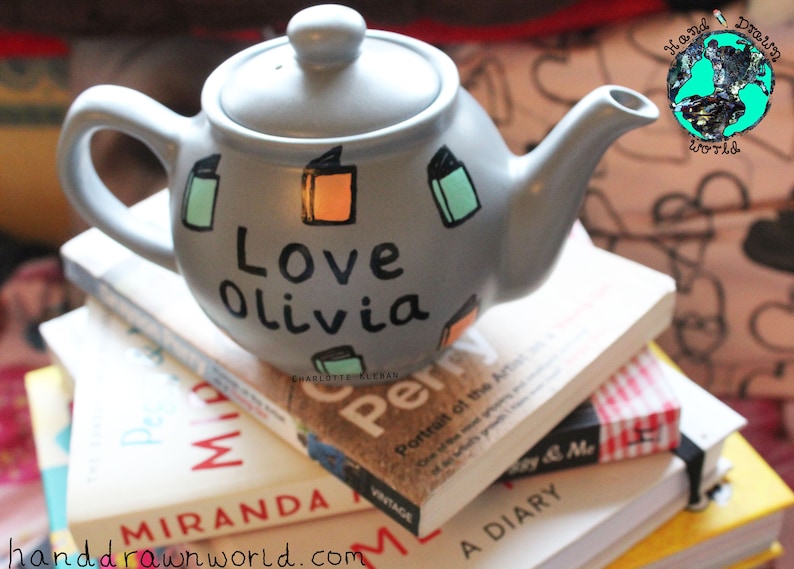 May include: A grey teapot with a white lid and black lettering that says "Love Olivia". The teapot is decorated with colourful book illustrations. The teapot is sitting on a stack of books.