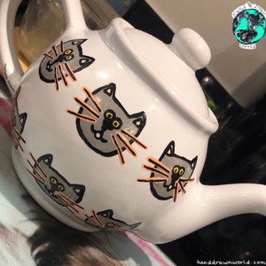 Op de afbeelding: Witte theepot met een handgeschilderd ontwerp van grijze katten met oranje snorharen. De theepot heeft een deksel en een tuit. De tekst "Cat's Whiskers" staat op de zijkant van de theepot.