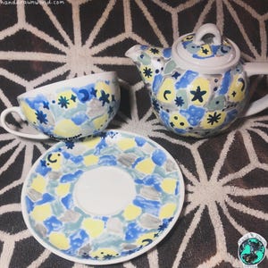 May include: A hand-painted ceramic tea set comprising a teapot, teacup, and saucer. The set is adorned with blue, yellow, and grey abstract shapes and star patterns. The teacup and saucer bear the artist's signature.
