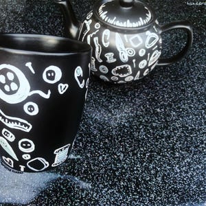 May include: A black ceramic mug and teapot set decorated with white illustrations of sewing notions. The mug features a handle and is covered in images of buttons, measuring tapes, and other sewing-related items. The teapot has a matching design.