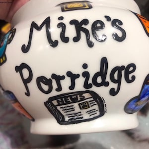 May include: White ceramic bowl with black lettering that reads "Mike's Porridge". The bowl is decorated with colorful drawings of a newspaper, a cup of coffee, and other objects.