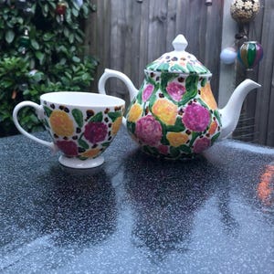 May include: A white ceramic teapot with a matching teacup, both hand-painted with vibrant yellow and pink floral patterns, green leaves, and small brown accents. The teapot features a lid with a pointed finial and a curved handle and spout.