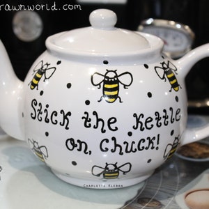 May include: White ceramic teapot with a black and yellow bee design and the text "Stick the Kettle on, chuck!"  The teapot has a black handle and a black lid.